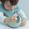 子どもがディンキーカップを持ちながらグレーの吸盤ボウルで自分で食事をしている写真