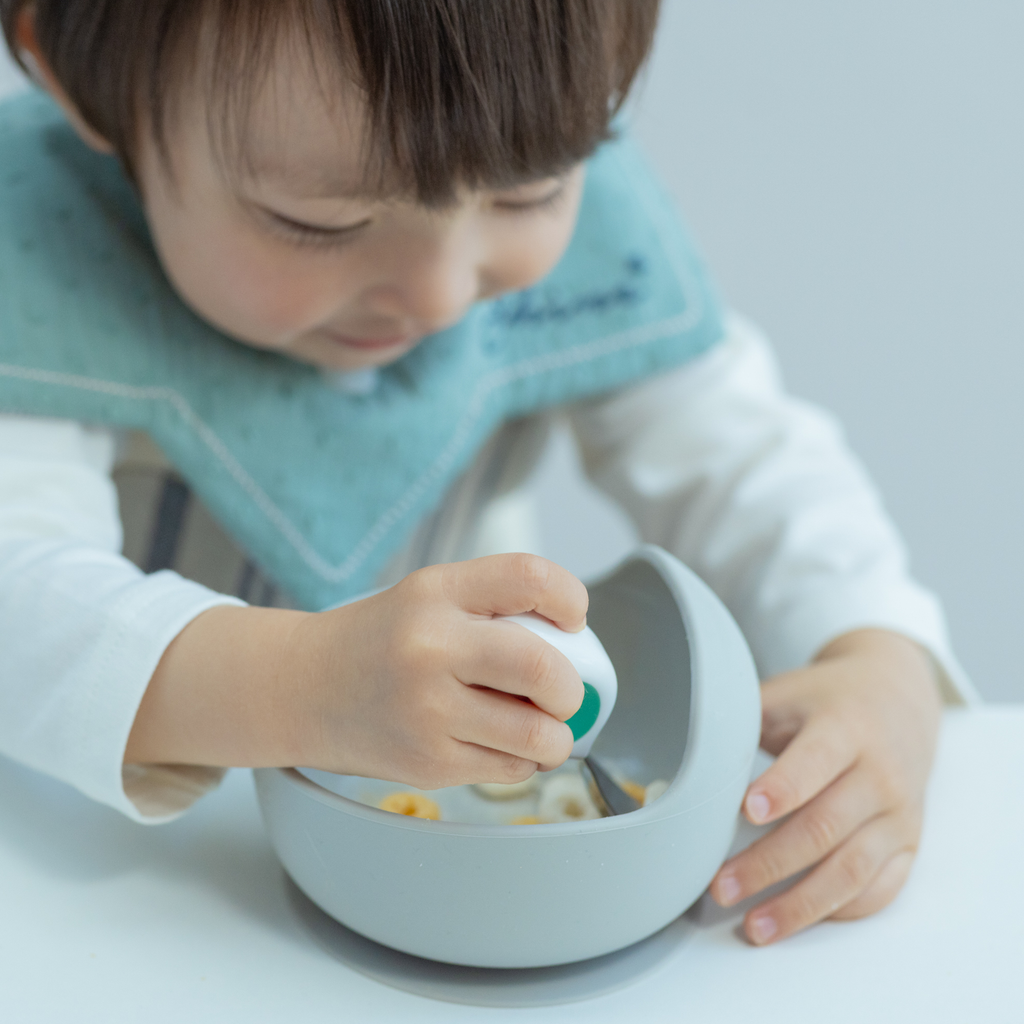 子どもがディンキーカップを持ちながらグレーの吸盤ボウルで自分で食事をしている写真